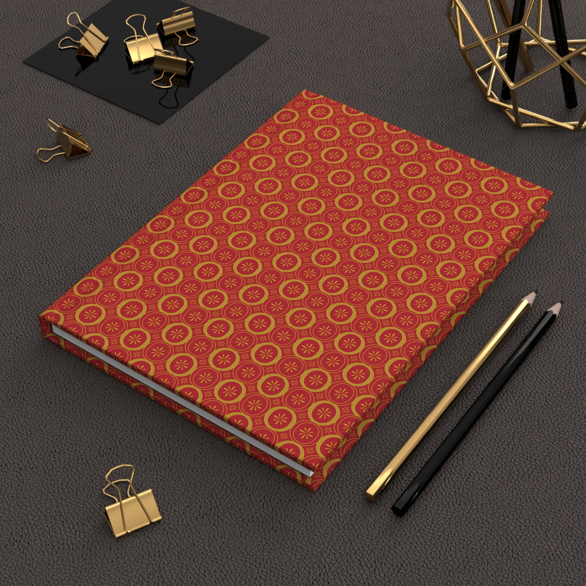 Red notebook with gold pattern on a dark surface with office supplies.